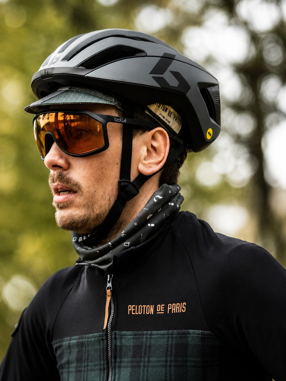 Person wearing a black helmet and sunglasses with 'Peloton de Paris' jacket outdoors.