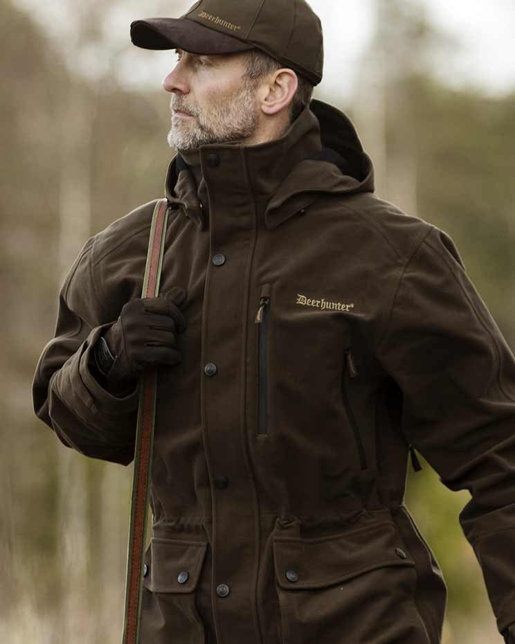 Man wearing a brown hunting jacket and cap with 'Deerhunter' branding, holding a rifle, in a forest setting.