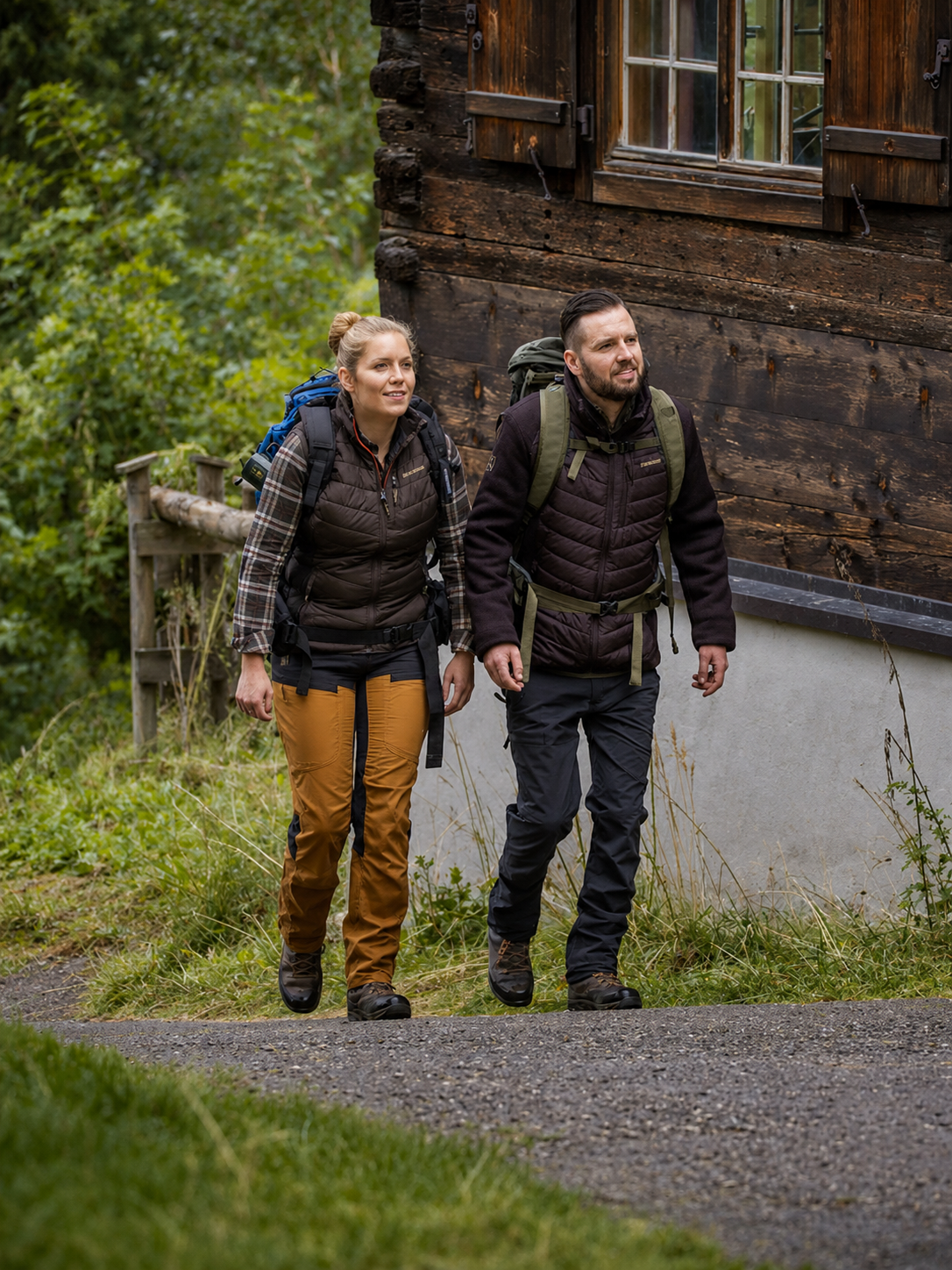 Two hikers with backpacks walking on a path near a wooden building.