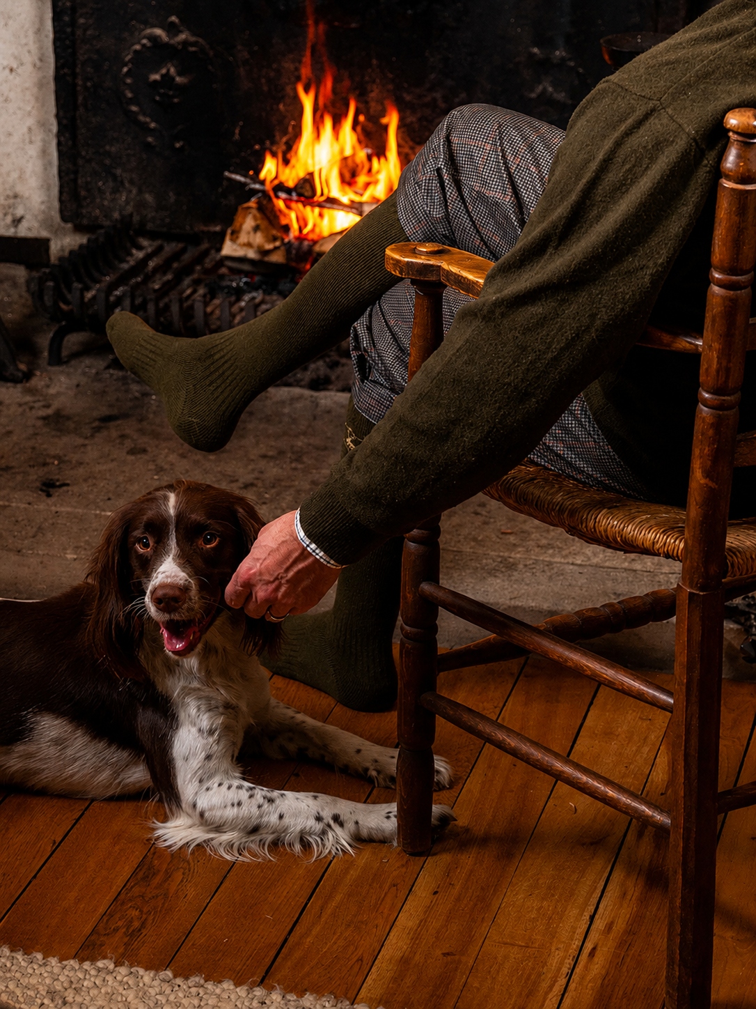 Person sitting in a chair with a dog on a wooden floor in front of a fireplace.