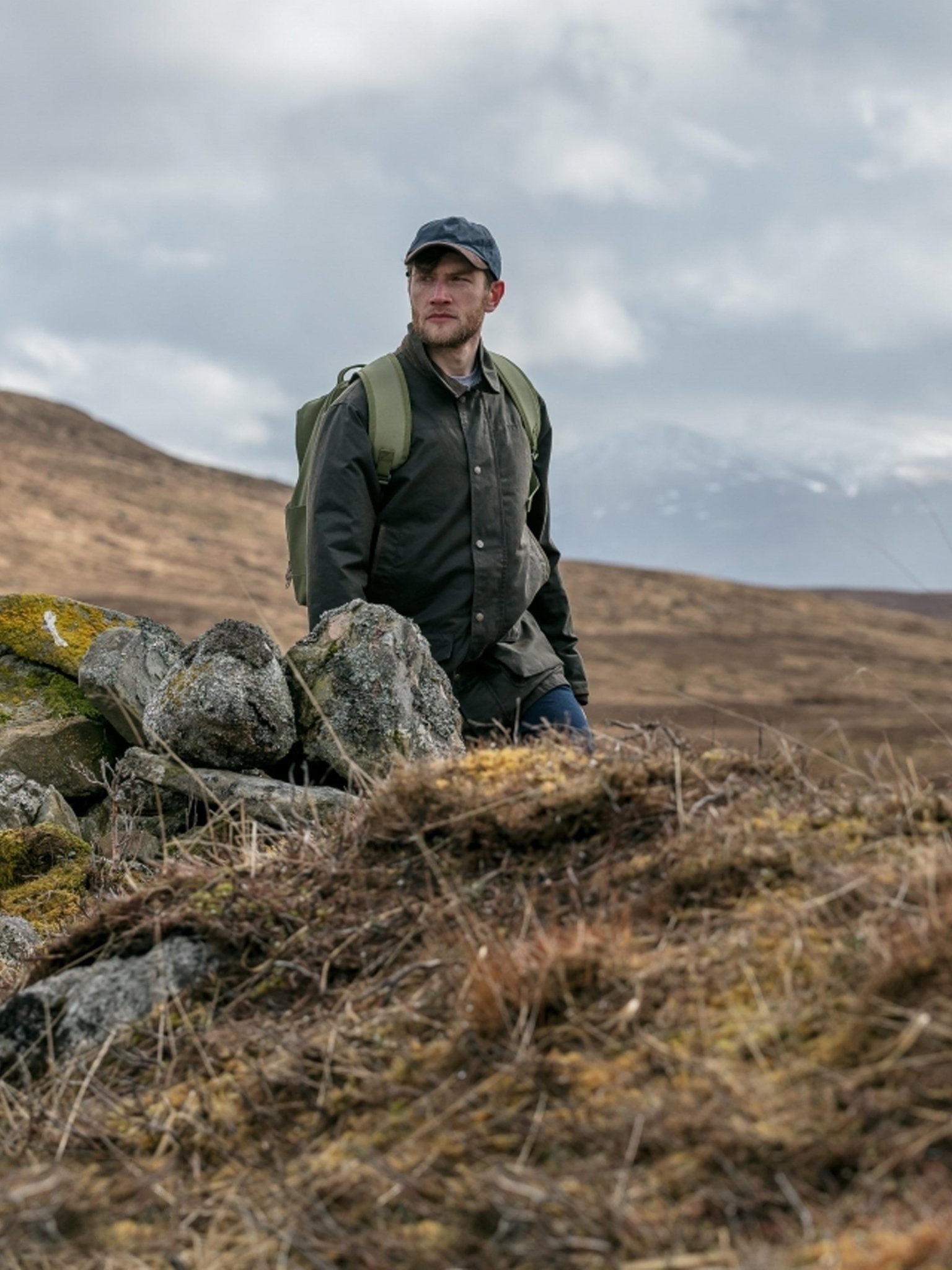 Man standing on a rocky hillside with a backpack, wearing outdoor gear.