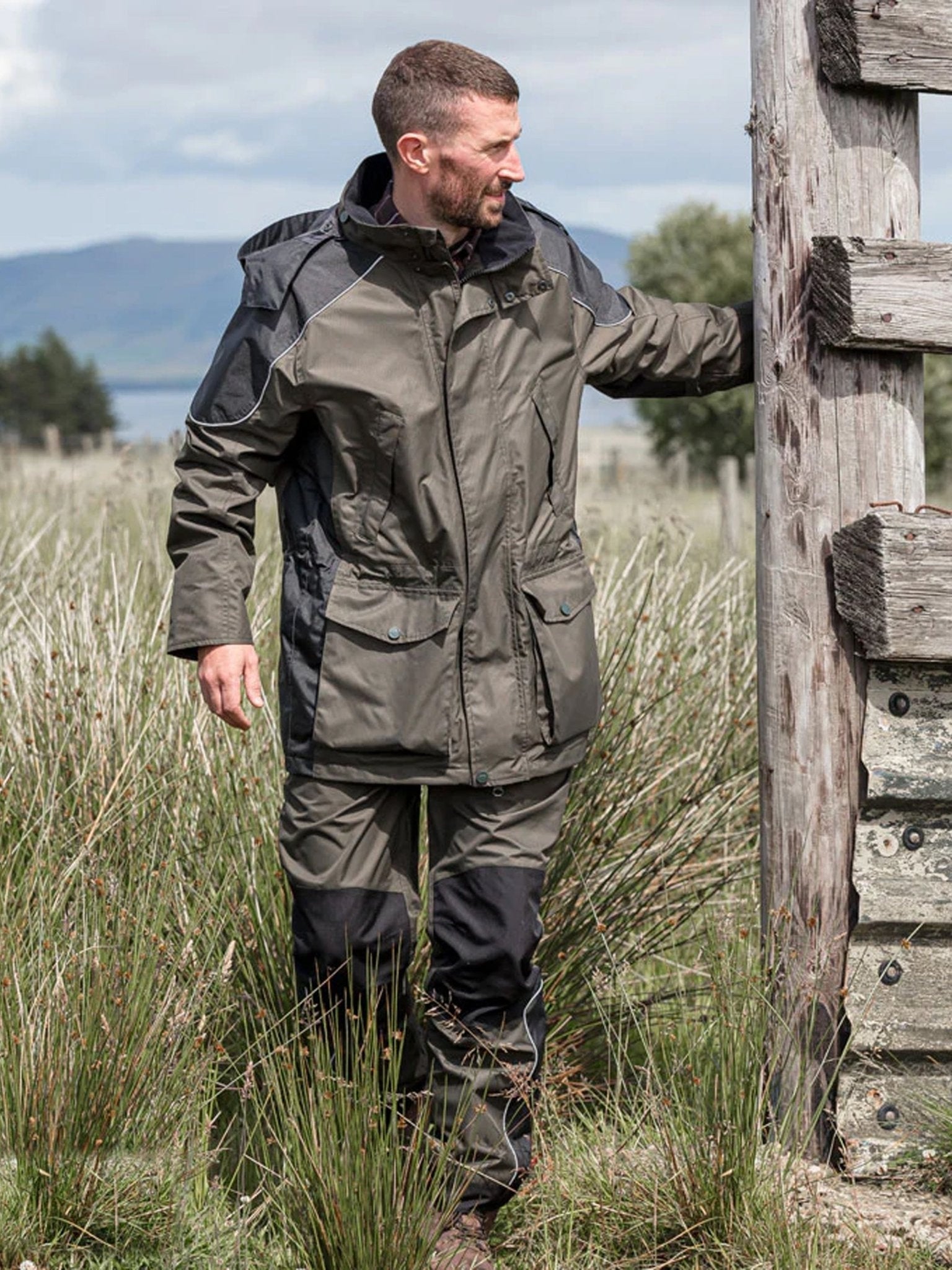 Man wearing a green and black jacket and pants standing in a field with mountains in the background.