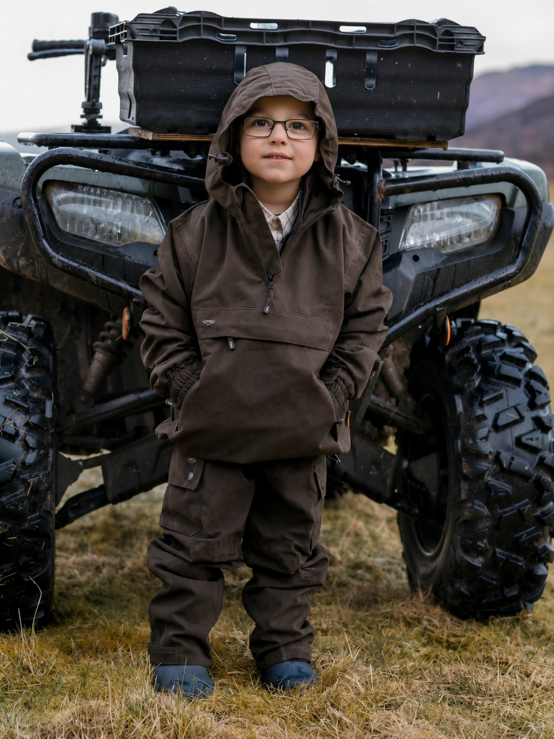 Child in brown jacket and pants standing in front of a large black ATV on a grassy field.