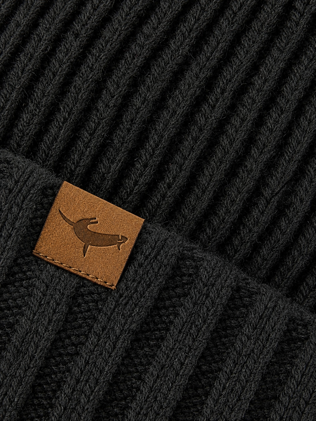 Close-up of a black knit beanie with a brown leather tag featuring a logo.