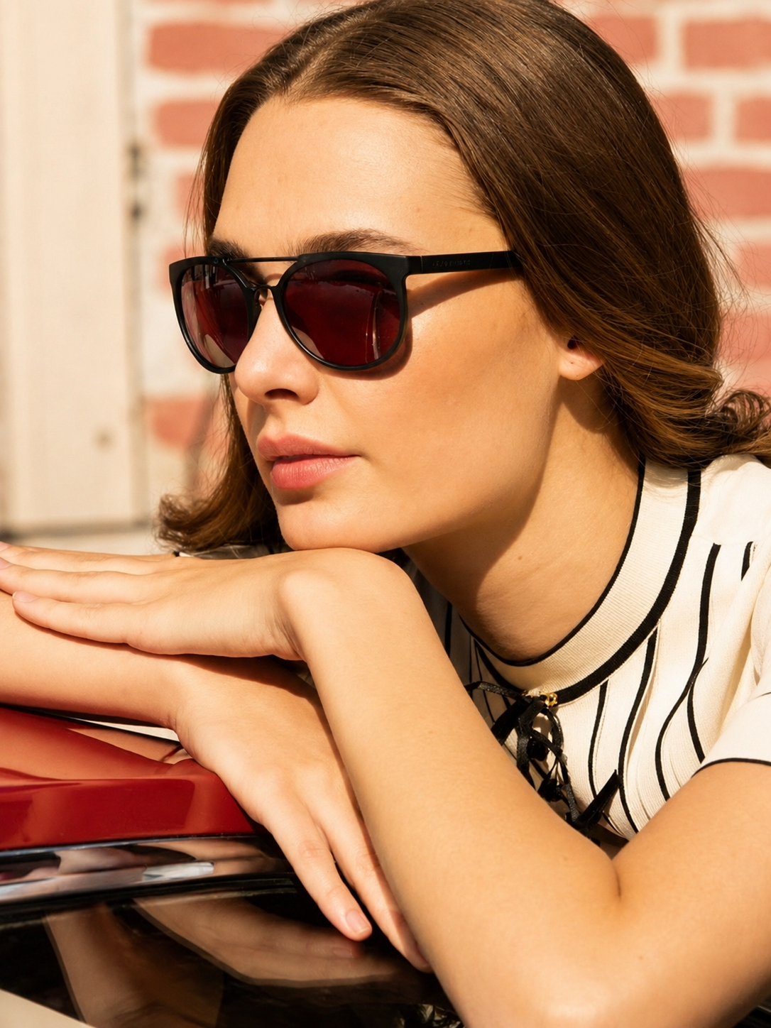 Woman wearing sunglasses leaning on a car with a brick wall background