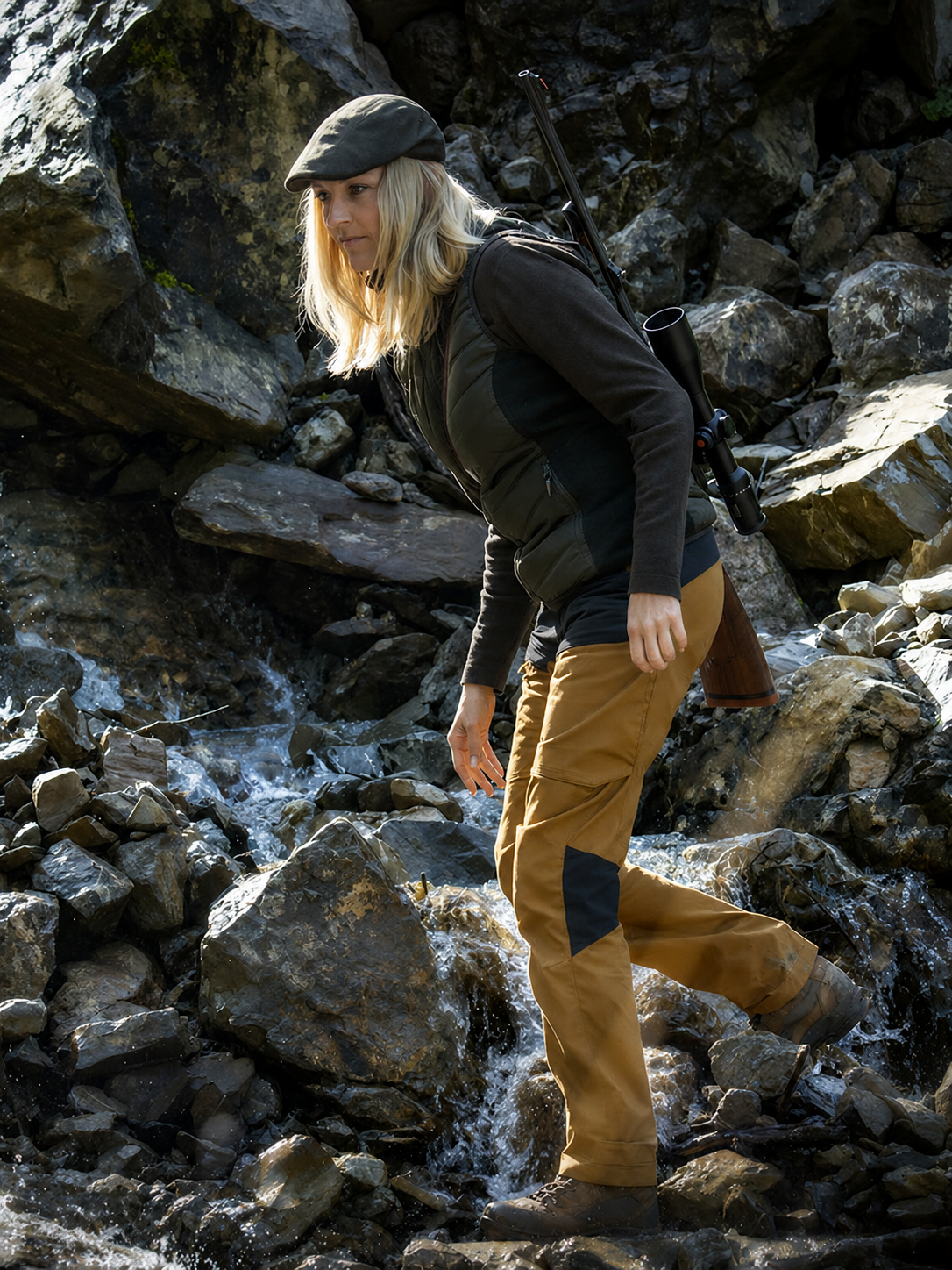 Person hiking through a rocky stream wearing a cap, jacket, and pants.