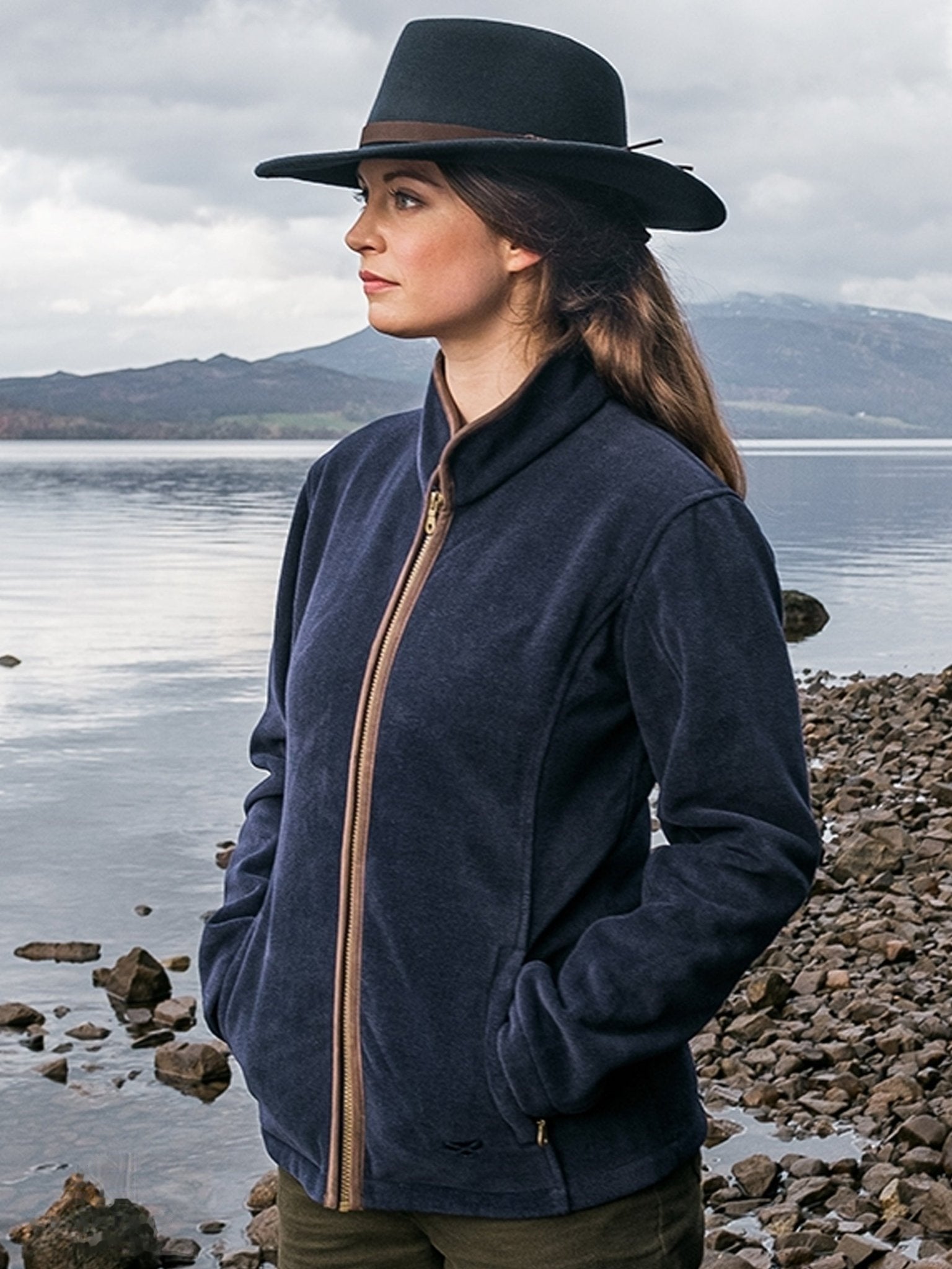 Woman wearing a navy jacket and hat by a lakeside with mountains in the background