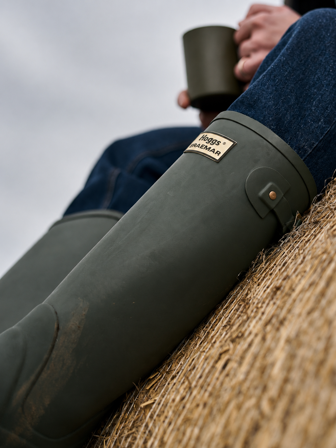 Person wearing green wellington boots with a visible Hoggs of Fife logo, sitting on a hay bale.