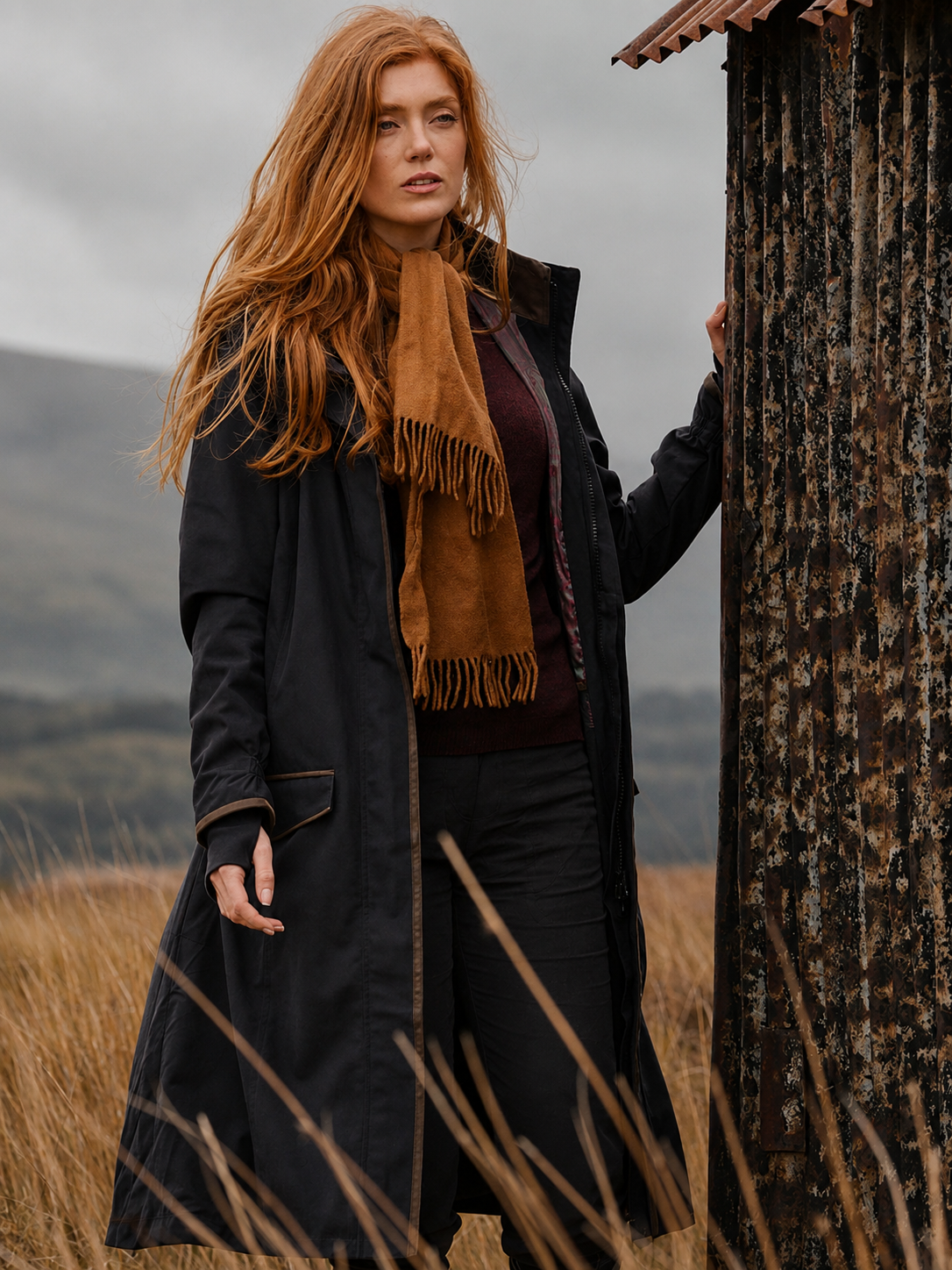 Woman in a long dark coat and brown scarf standing in a field with mountains in the background
