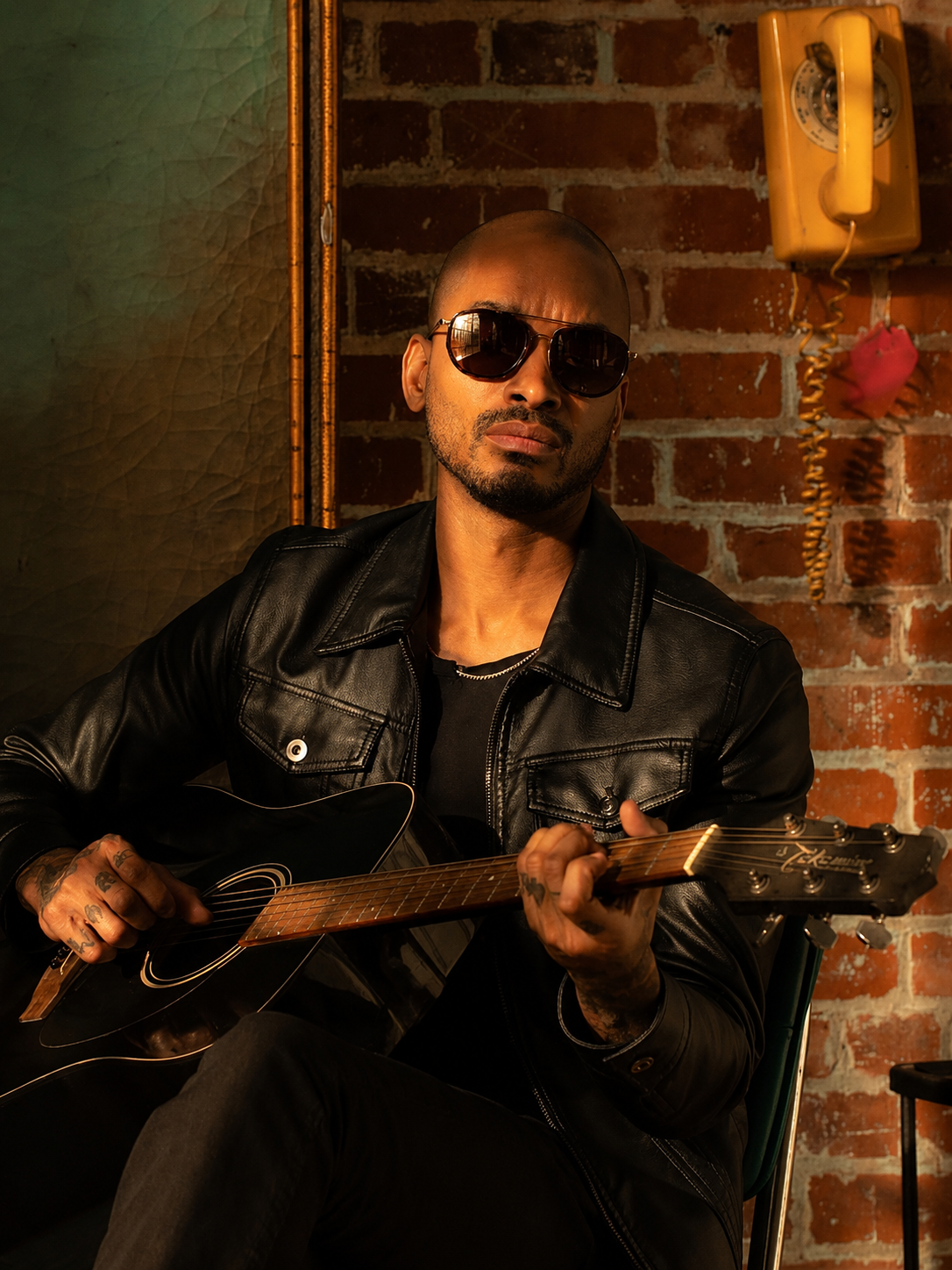Man playing guitar against a brick wall with a vintage phone and heart decoration wearing Serengeti sunglasses.